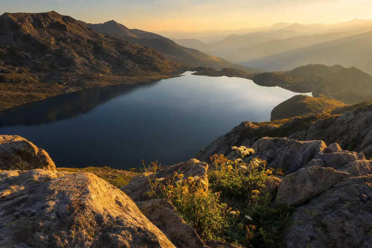 A dramatic mountain landscape at golden hour with a sharp rocky foreground, a still lake, and layered peaks fading into the distance