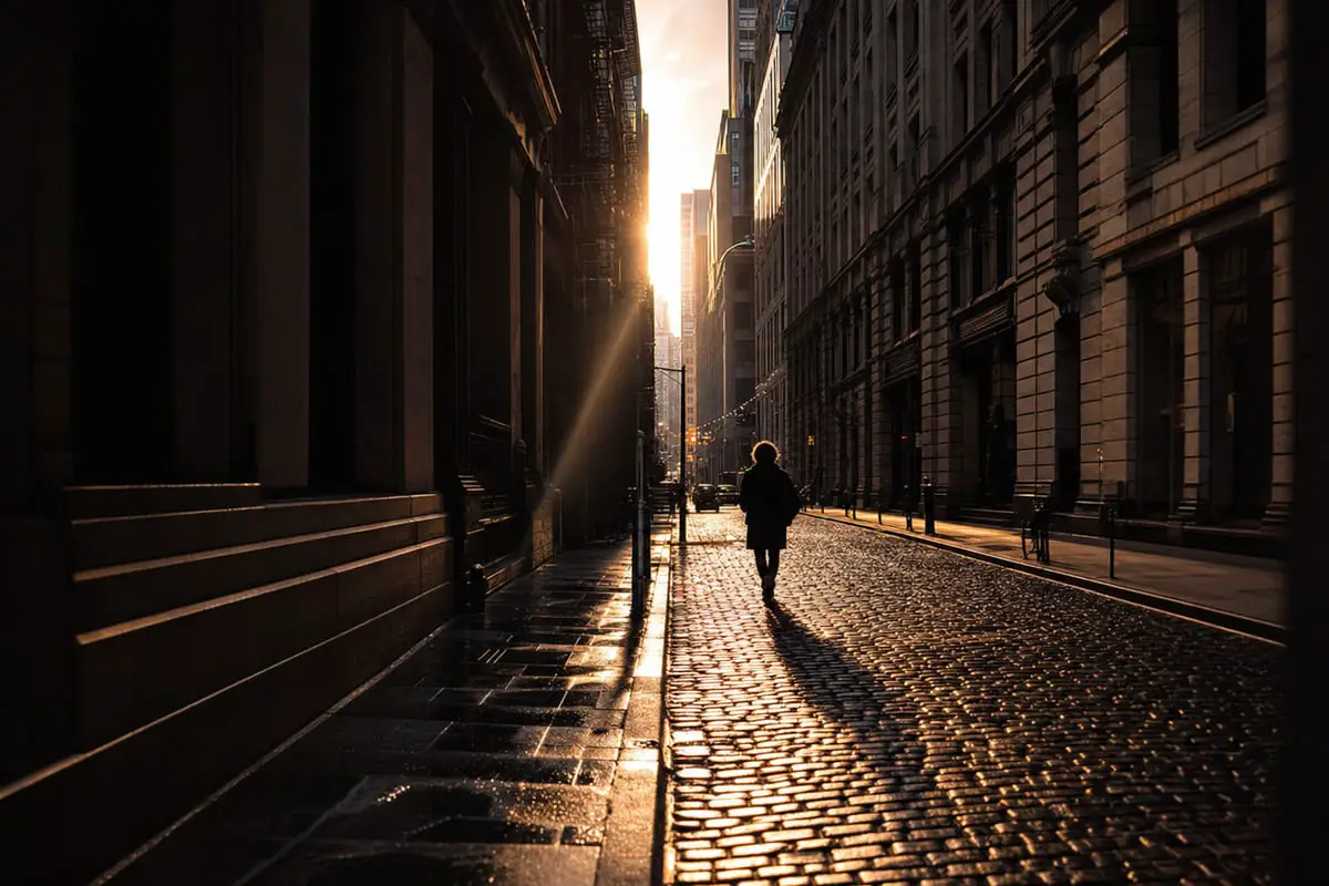 A candid black-and-white street photography scene of a person walking through a beam of light between city buildings